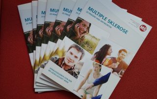 7 Ausgaben der Broschüre" Multiple Sklerose: 25 Fragen und Antworten" liegen auf einem roten Stoff. Herausgegeben von der Multiple Sklerose Gesellschaft Wien im November 2020